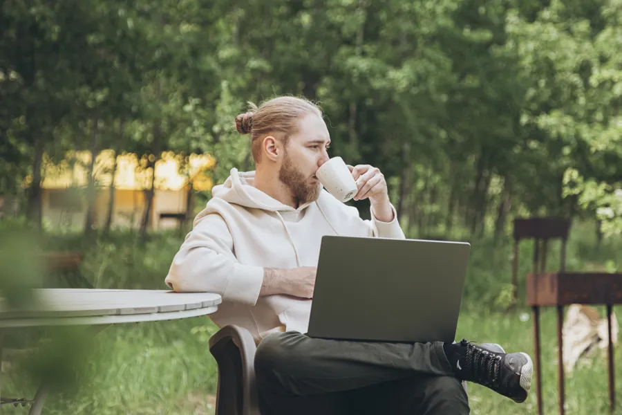 Ung mann med PC i fanget og kaffekopp, ute i et hagemiljø.