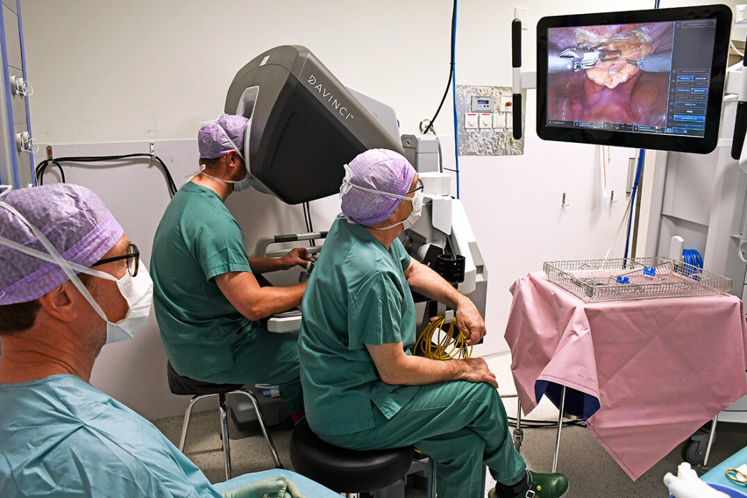 En person styrer en operasjonsrobot, to andre følger med på skjerm