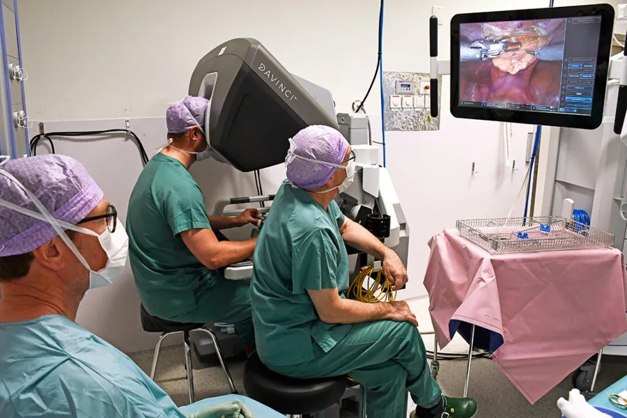 En person styrer en operasjonsrobot, to andre følger med på skjerm