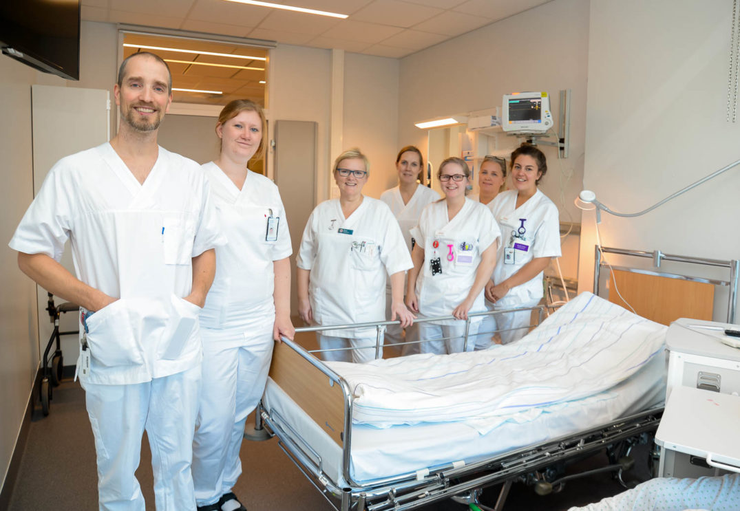 En gruppe mennesker i hvite laboratoriefrakker står ved siden av en seng