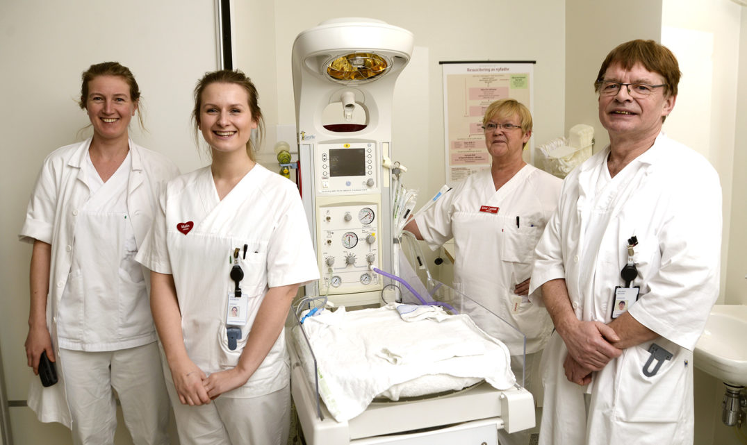 En gruppe mennesker i hvite laboratoriefrakker står ved siden av en sykehusseng