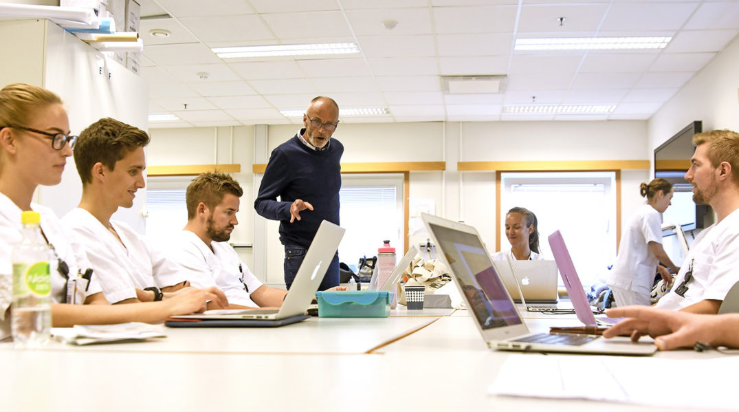 En person som snakker til en gruppe mennesker som sitter ved et bord med bærbare datamaskiner