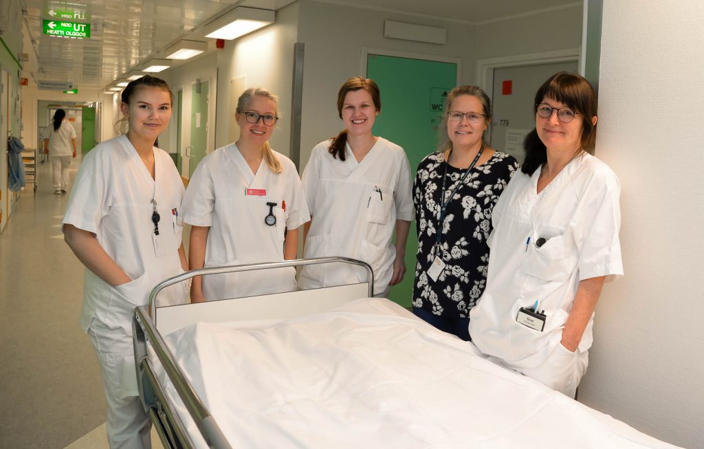 En gruppe kvinner i hvite laboratoriefrakker står ved siden av en sykehusseng