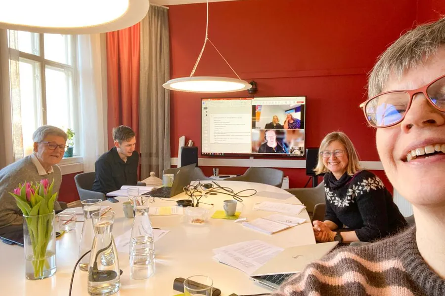 Fire mennesker som sitter rundt et bord. Tre mennesker deltar også via dataskjerm