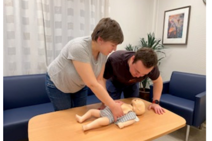 Voksen kvinne og mann øver med babydokke på et stuebord.