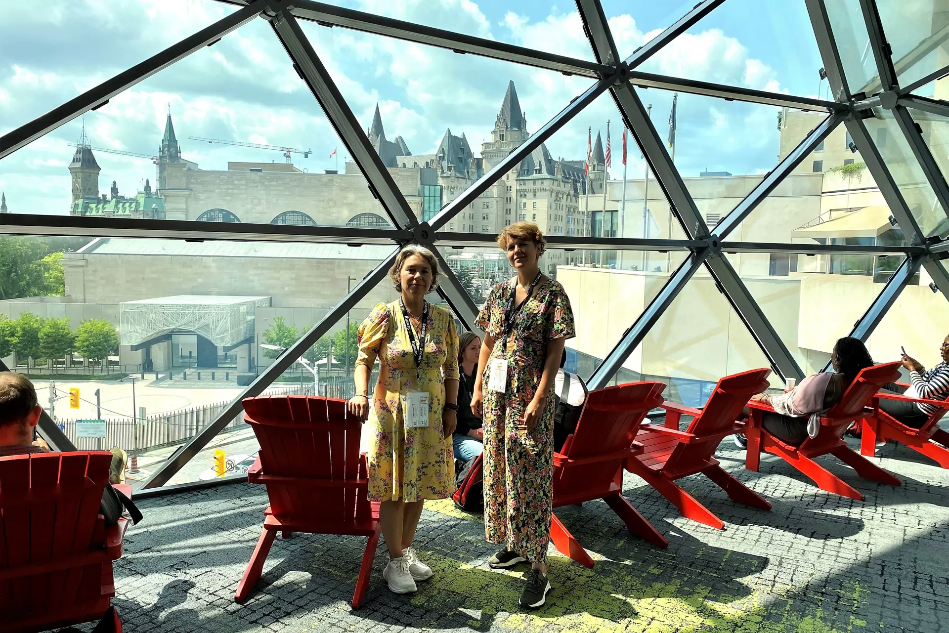 Et par kvinner som står foran et metallgjerde med røde stoler
