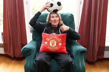 Døvblind dame sitter i lenestol med fotball på hodet. Hun tuller og ler.