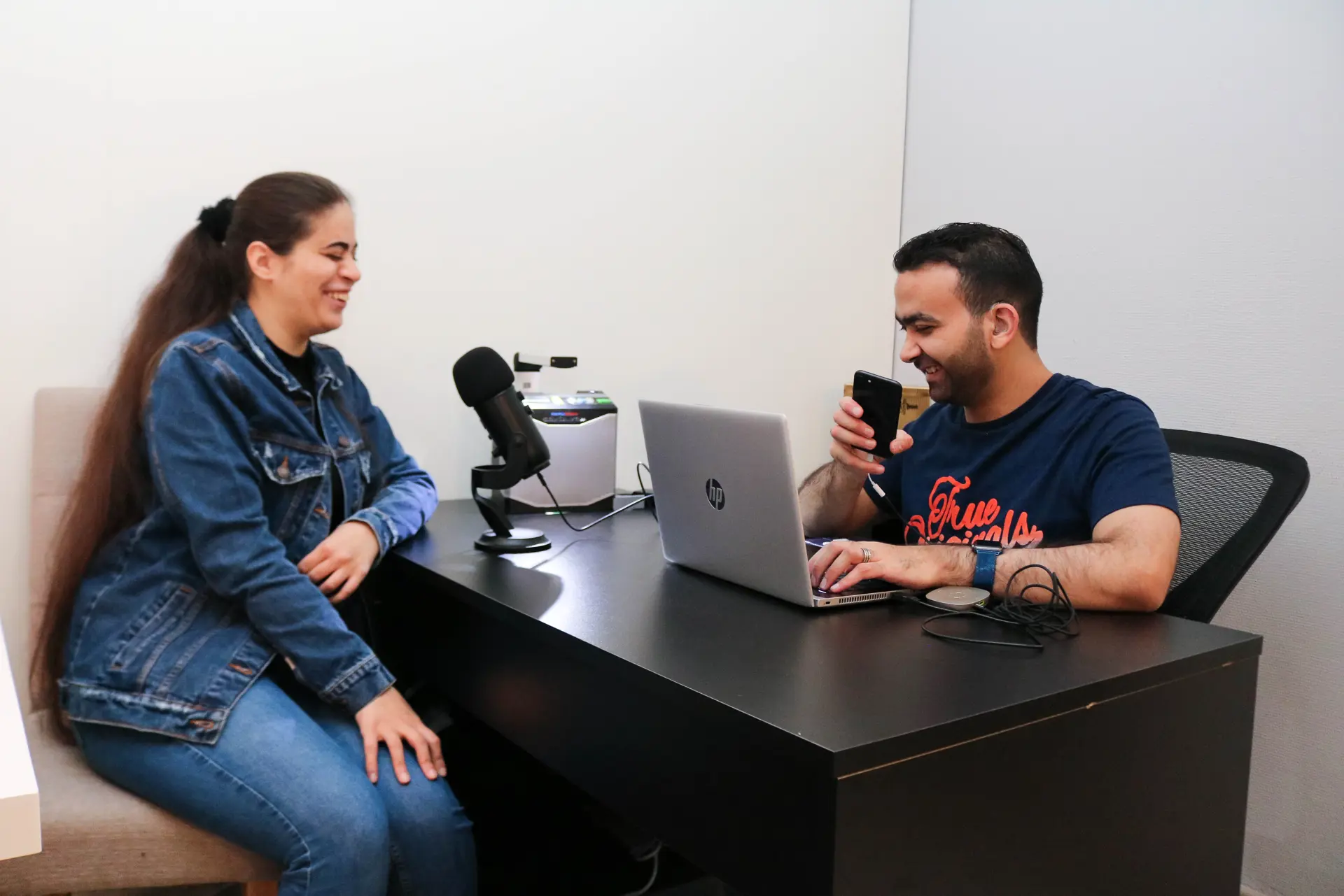 Kvinne og sitter på hver sin side av et bord med diverse podkast-utstyr og en datamaskin