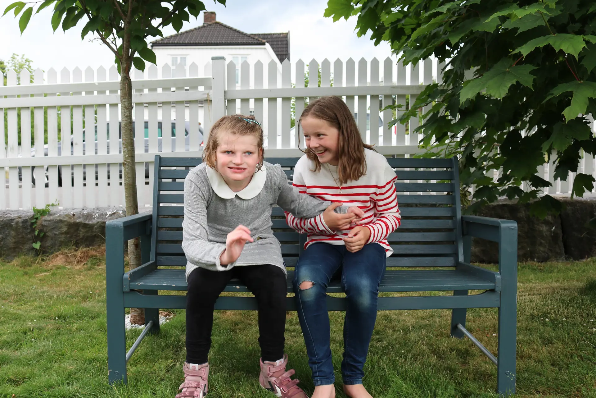 "To søstre i en sommerhage. Ei har rød og hvit-stripete genser og sitter på en benk, mens den andre går fra benken har på seg grå kjole.