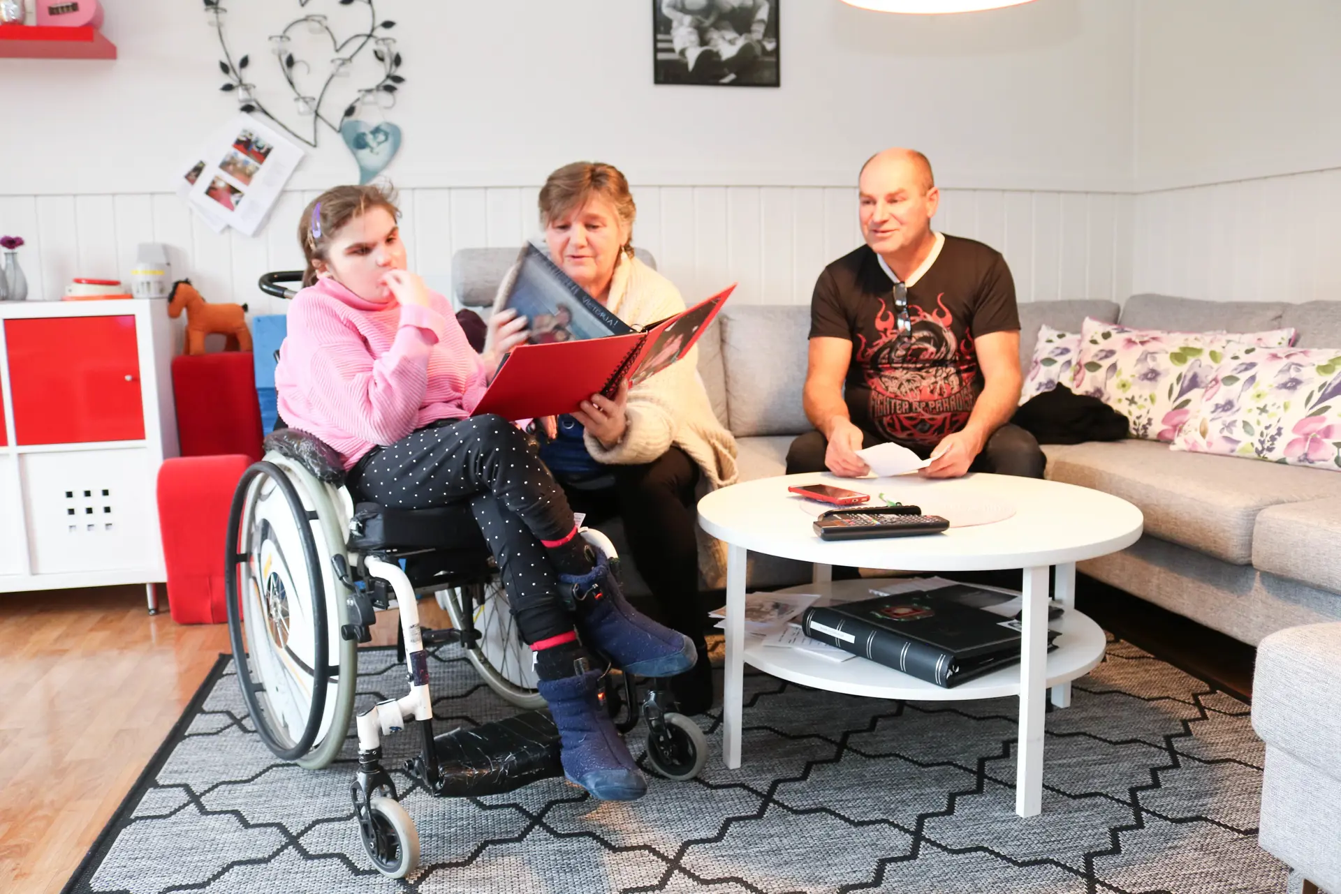 Jente i rullestol har besøk av sine foreldre. De sitter sammen rundt et stuebord og blar i gjennom et rødt fotoalbum