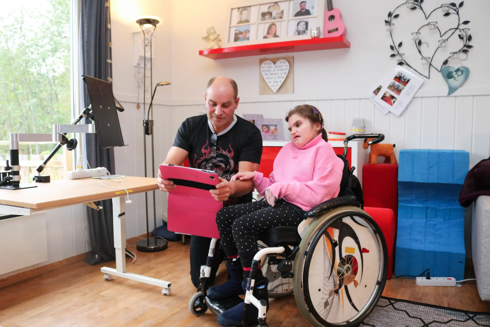 En mann står på kne ved siden av en kvinne i rullestol. Han holder i et rosa brett.