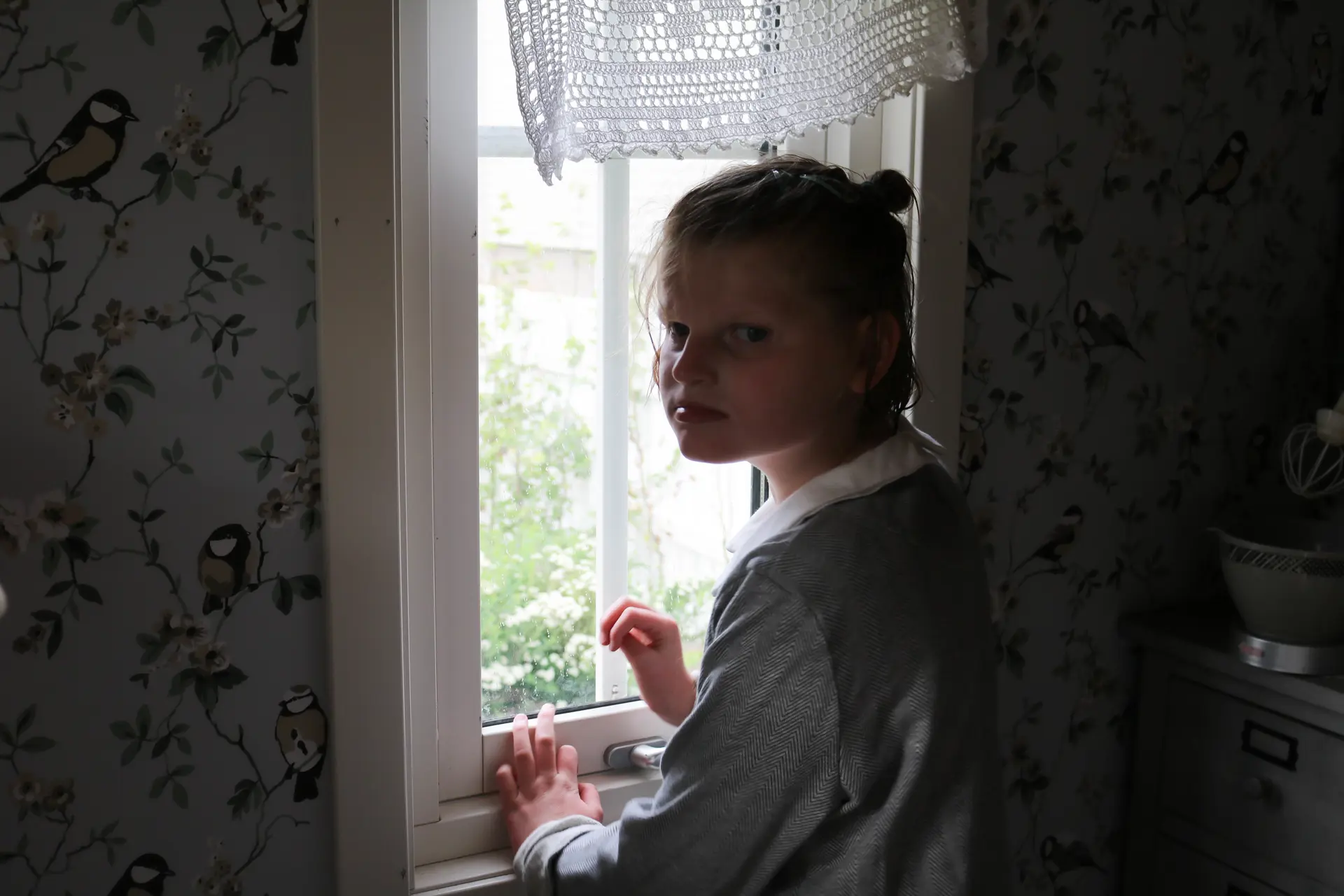 Et mørkt rom med ei jente som står med et smalt vindu, det er lyst utenfor