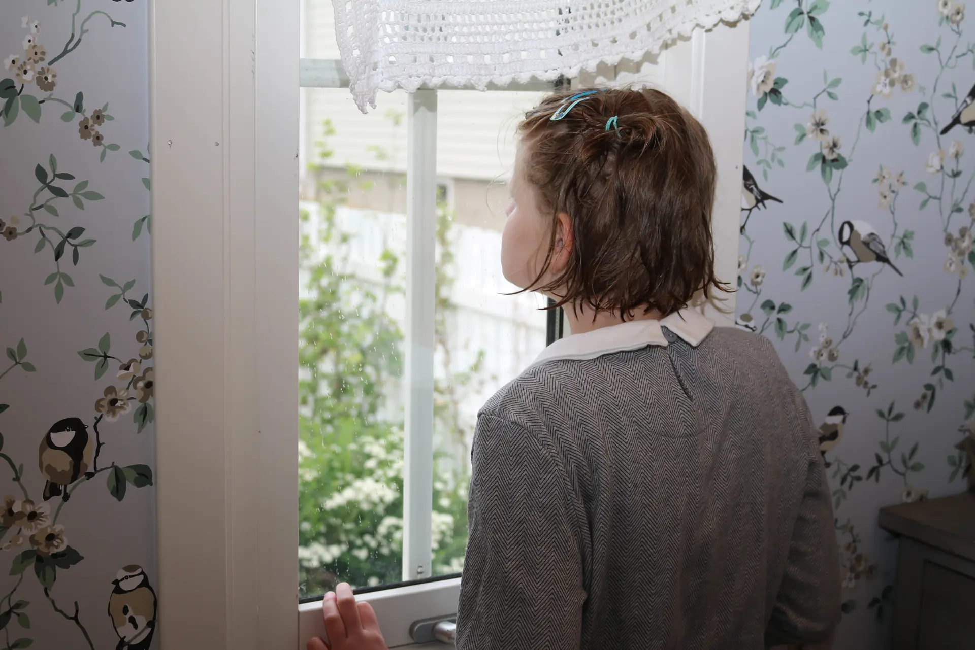Jente i grå kjole ser ut av et smalt vindu. Fuglemønster på tapet.