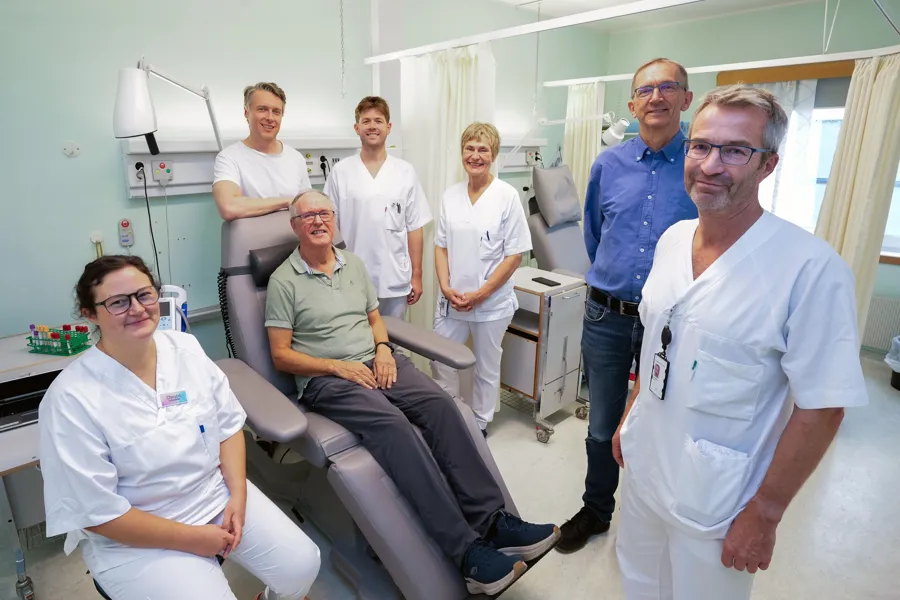 Gruppebilde: Fem hvitkledde ansatte som står, en sivilkledd mannlig pasient som sitter og en sivilkledd stående mann.