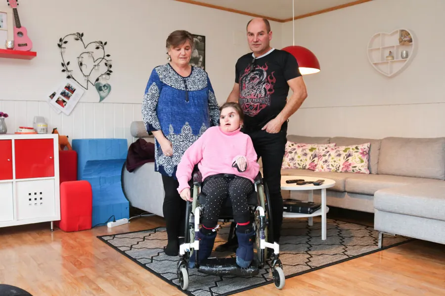 Voksen dame og mann sammen med en ung kvinne i rullestol. De er i en stue.