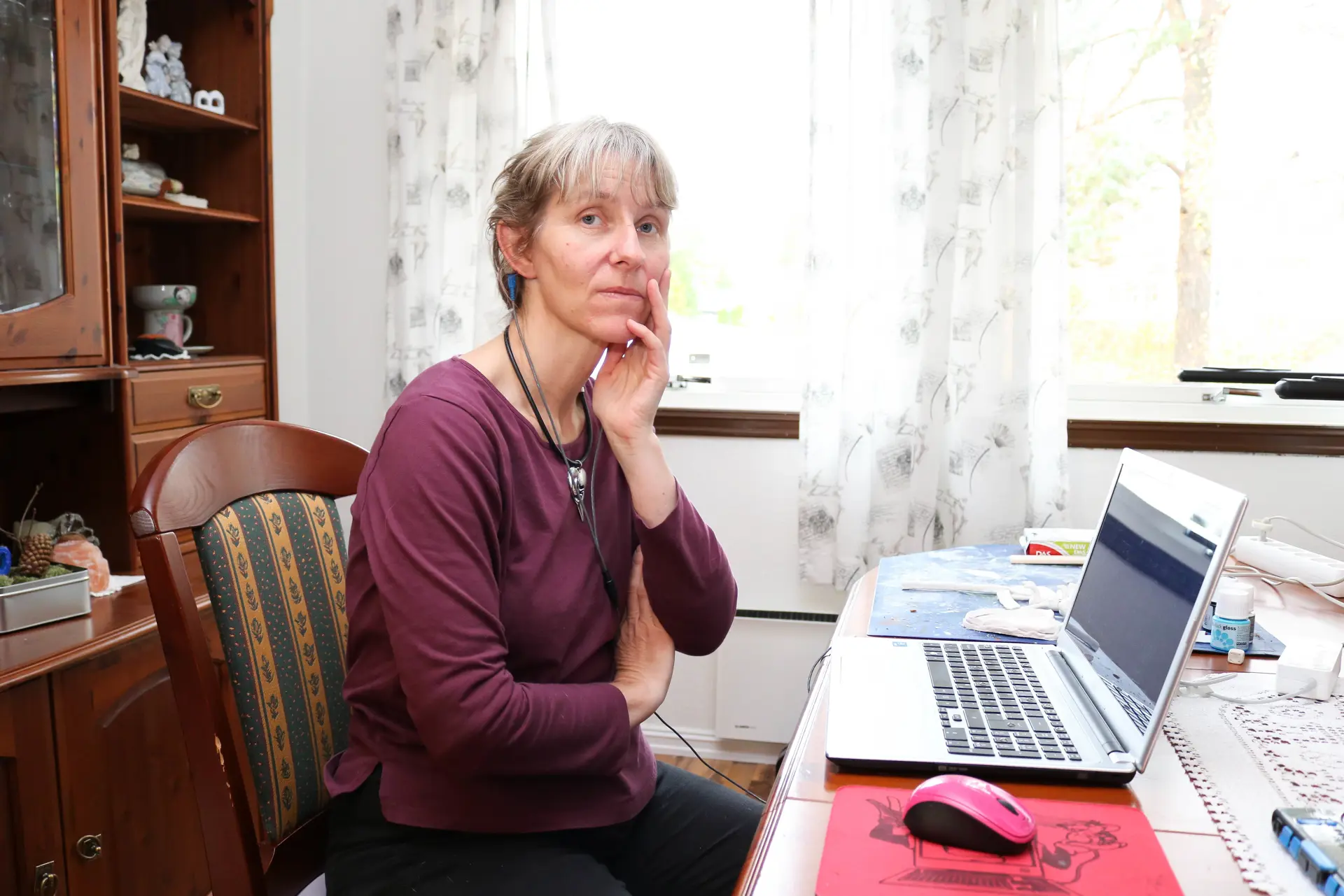 Spisestue. Kvinne i lilla genser sitter ved sin laptop og ser i kamera, hun holder venstre hånd mot kinnet sitt
