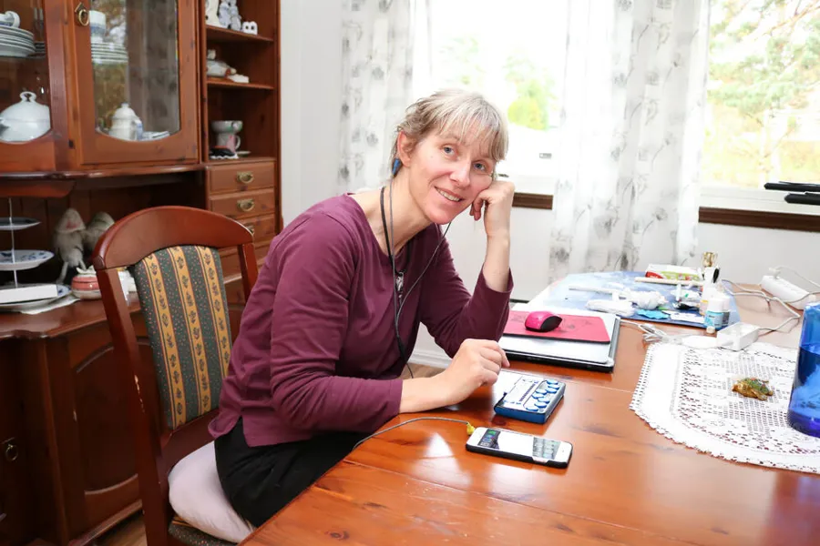 Kvinne med lilla genser i spisestue. Hun bruker sin mobil ved hjelp av headset og leselist. Hun hviler hodet på venstre hånd og smiler mot kamera.