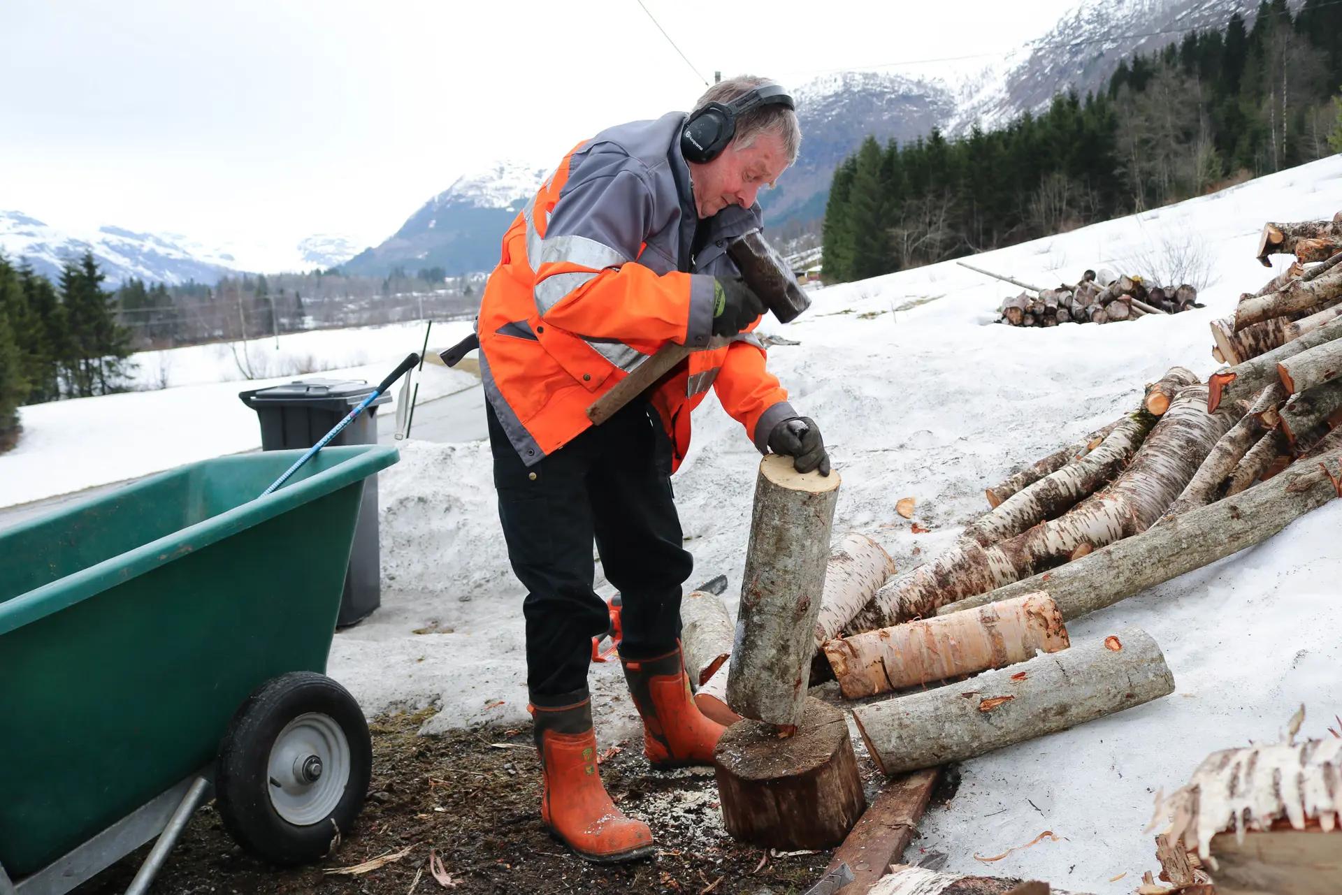 En mann  kløyver ved. Han er kledd i oransje jakke.