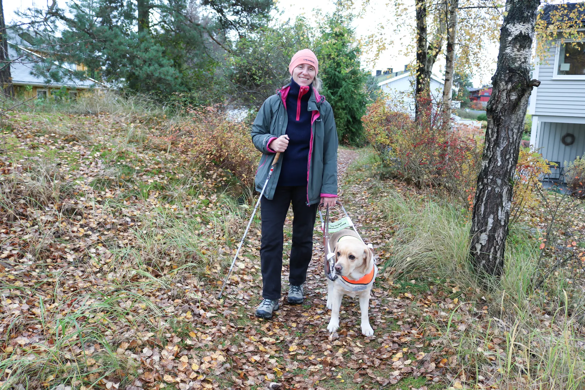 Høst i skogshage. Glad kvinne er ute og går med sin førerhund, i høyre hånd holder hun blindestokk. Flere hus kan skimtes i bakgrunnen.