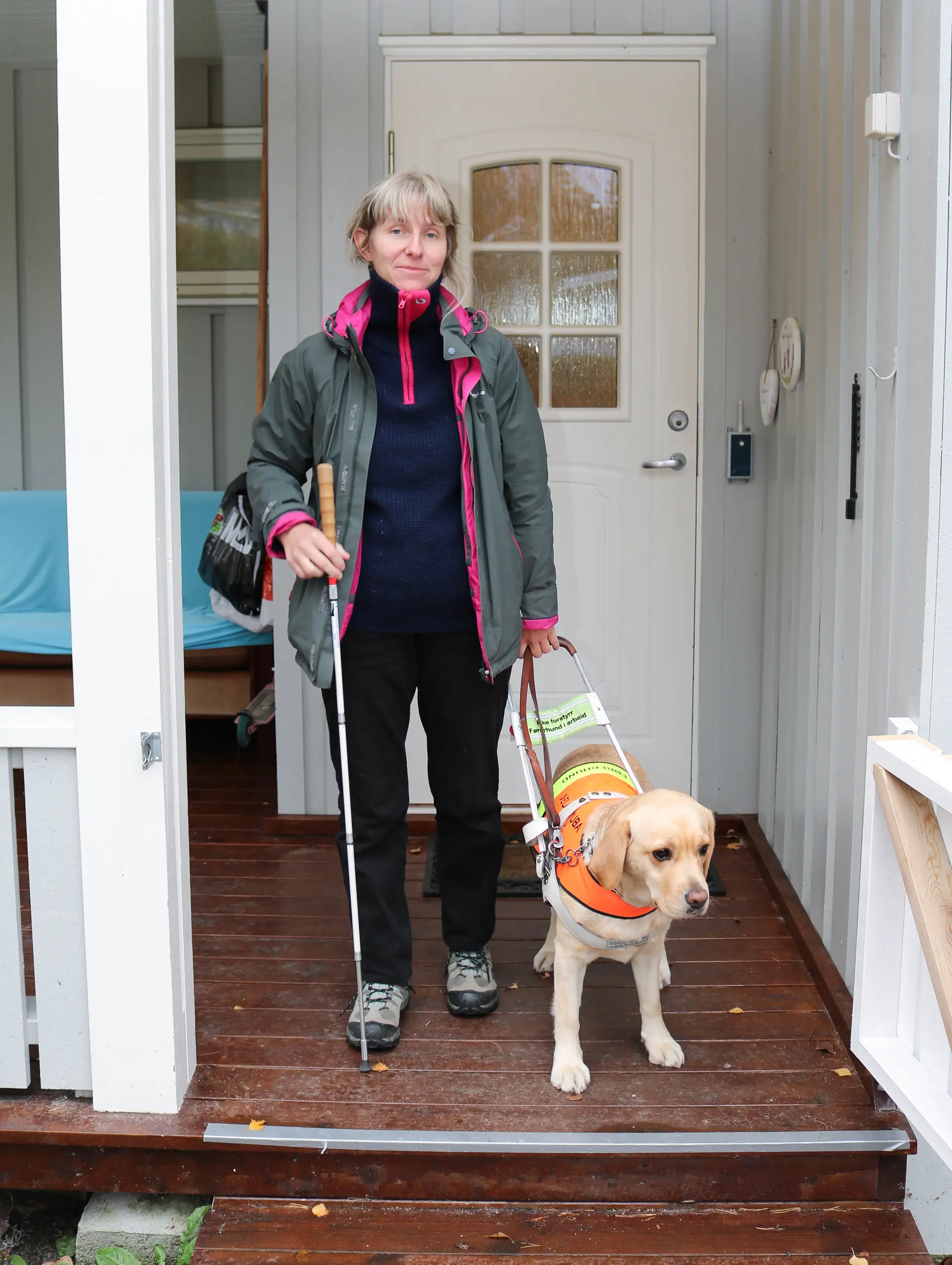 Foran ytterdør. Kvinne er på vei ut med sin førerhund, hun holder en blindestokk i høyrearm. Hun smiler til kamera