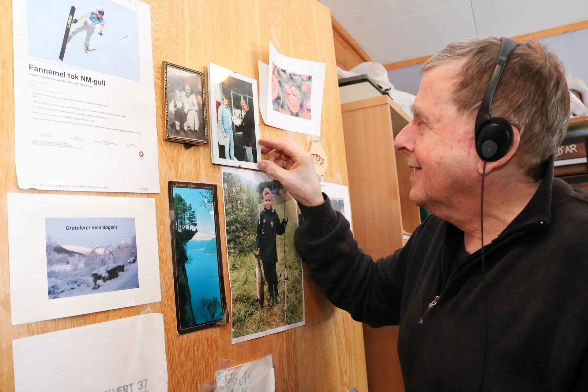 "Eldre mann med headsett viser fram en del bilder og utklipp som henger på døra til et hjemmekontor.