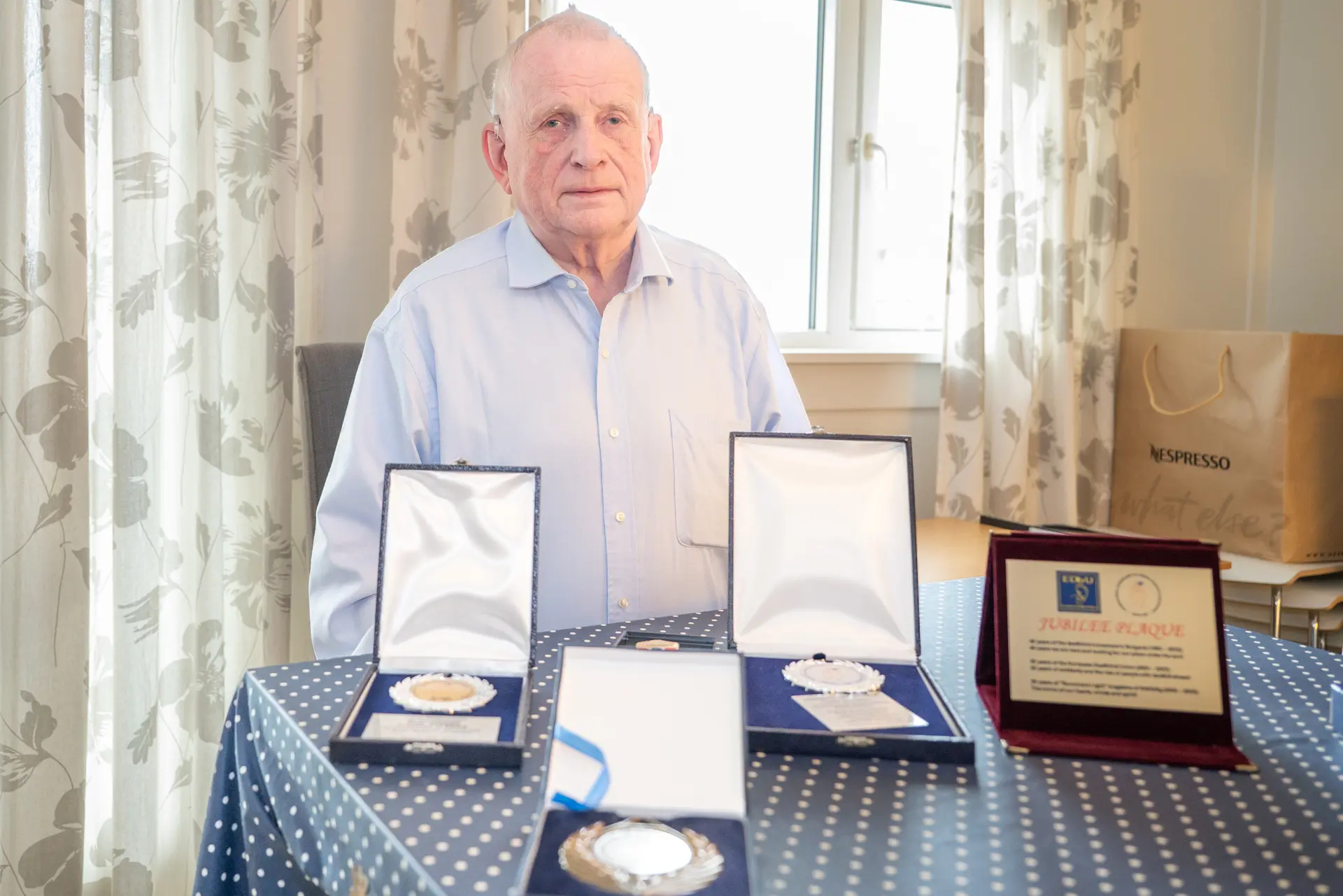 Eldre mann med blå skjorte sittende og på bordet foran ham er det plassert fire medaljer og et diplom.