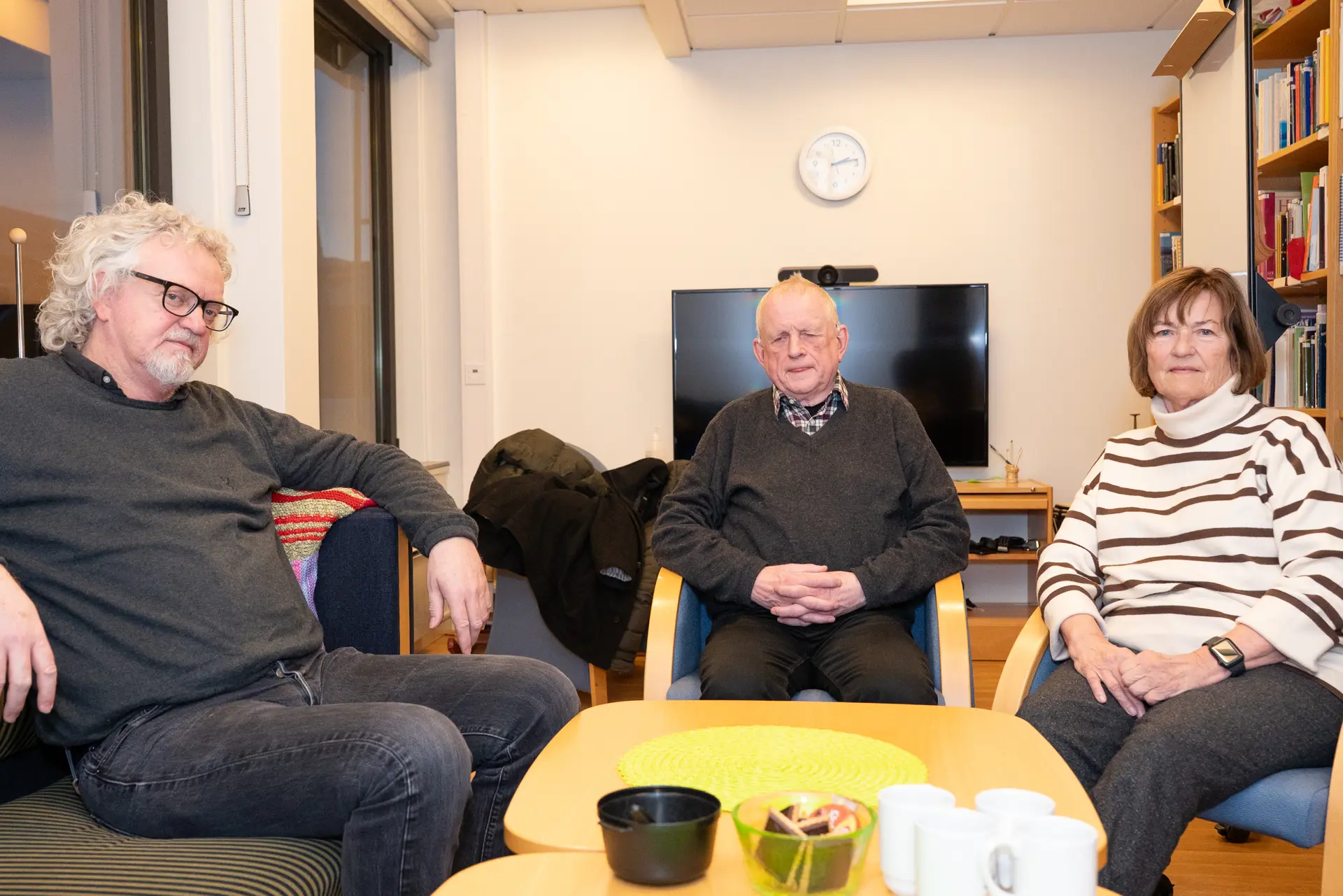 Tre personer sittende hvor alle ser mot kameraet. I bakgrunnen en TV-skjerm.