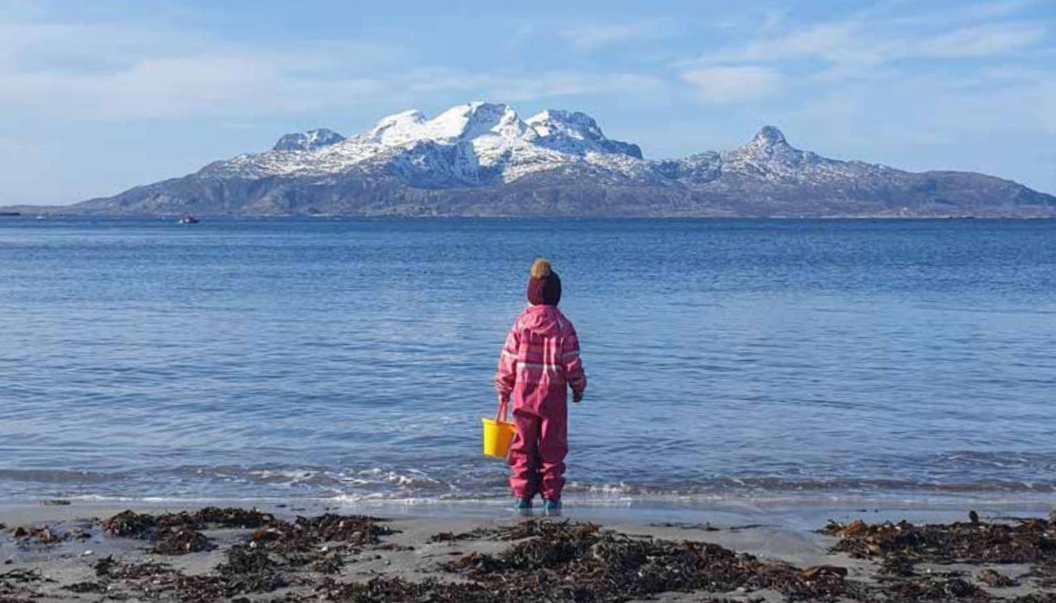 En jente i rosa klær som står på en strand med en bøtte. Hun ser mot fjorden og fjellene.