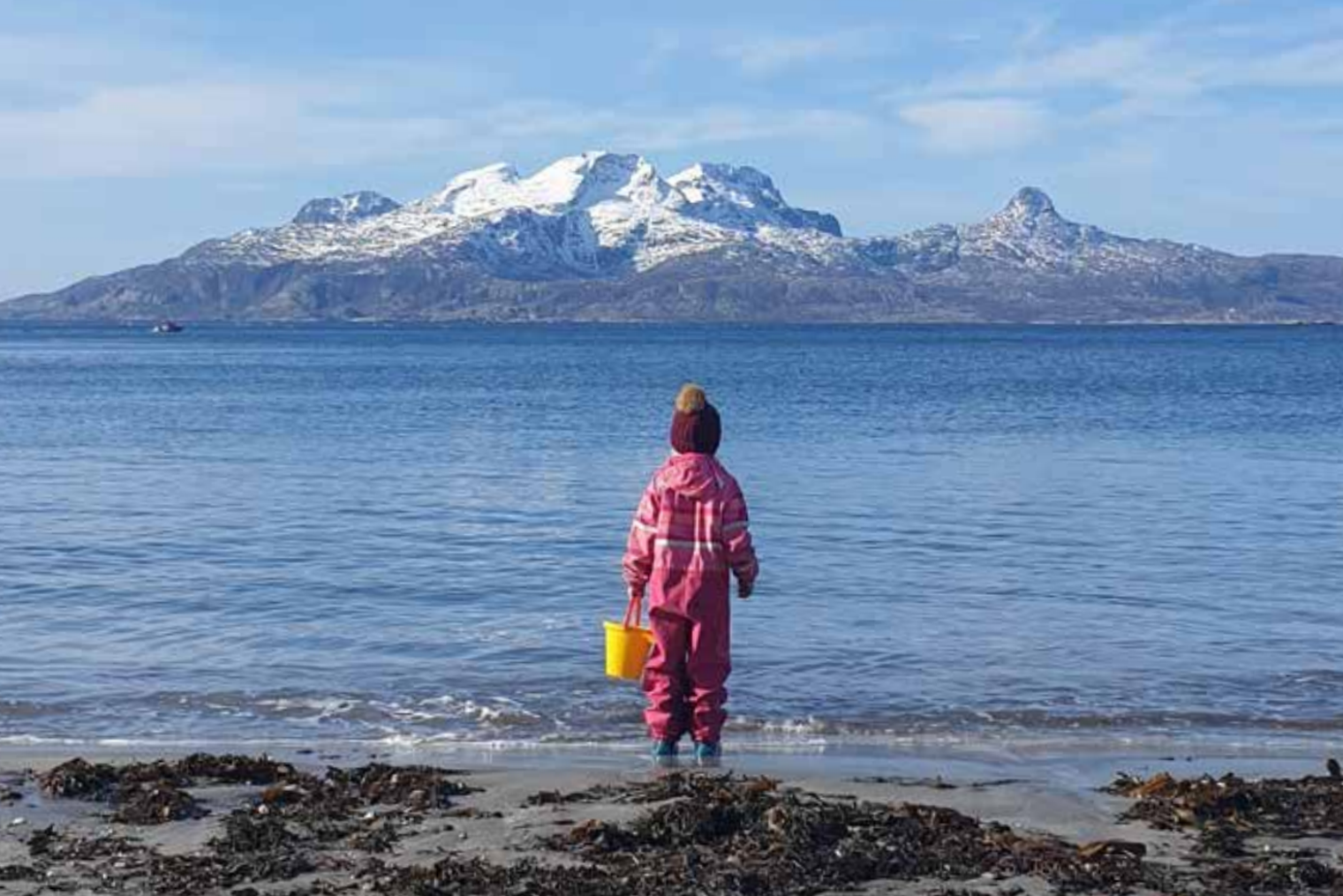 En jente i rosa klær som står på en strand med en bøtte. Hun ser mot fjorden og fjellene.